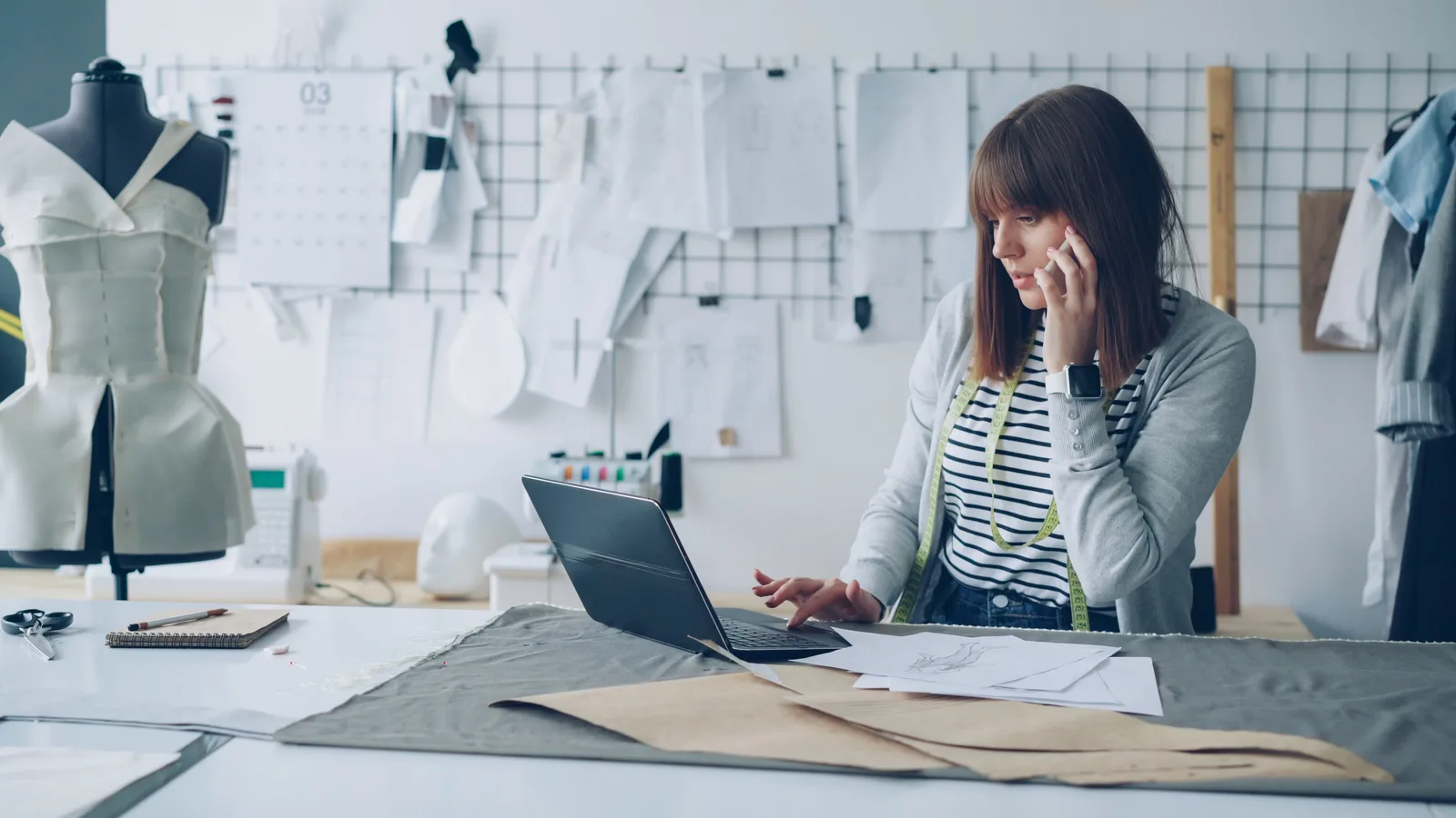 A female fashion designer working on her laptop in a creative design studio, surrounded by design sketches.